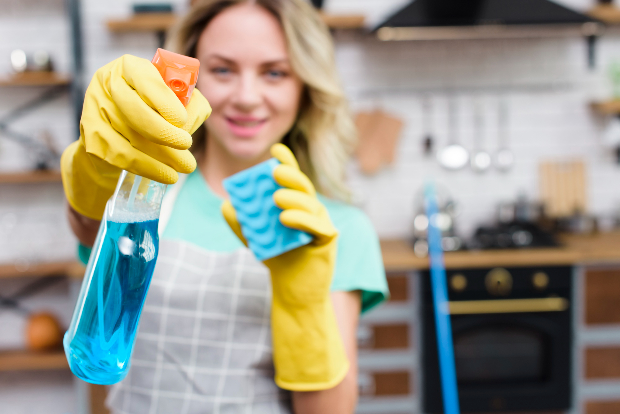 Woman wearing yellow gloves holding a bottle of cleaning solution and a sponge, implying housework or cleaning activity.